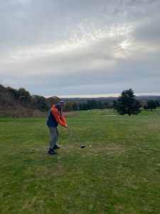 Brent Singer is in the distance getting ready to hit a golf ball with a golf club on a sunny day. 