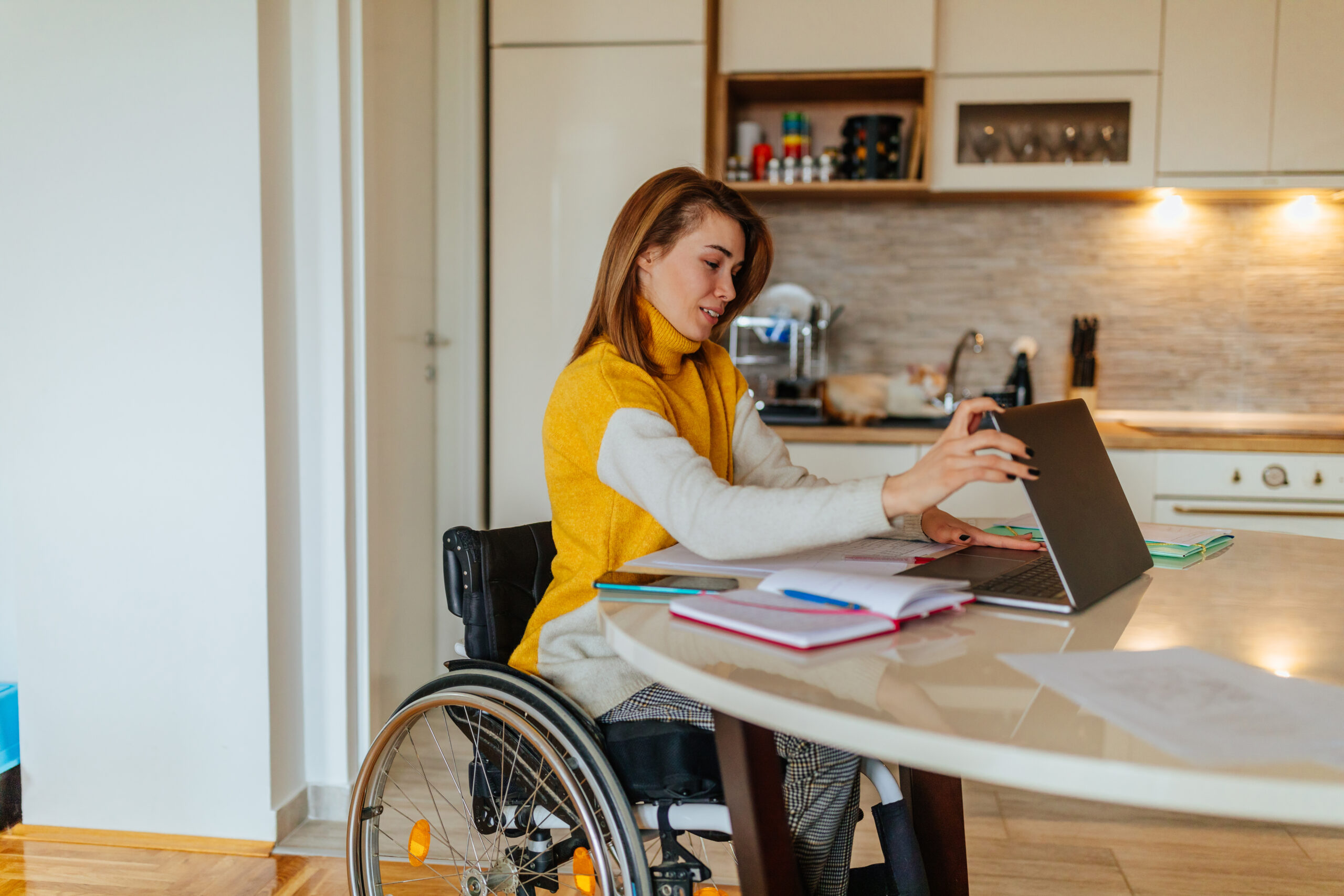 Woman in wheelchair sits at kitchen table with notebook and phone, and is opening laptop.